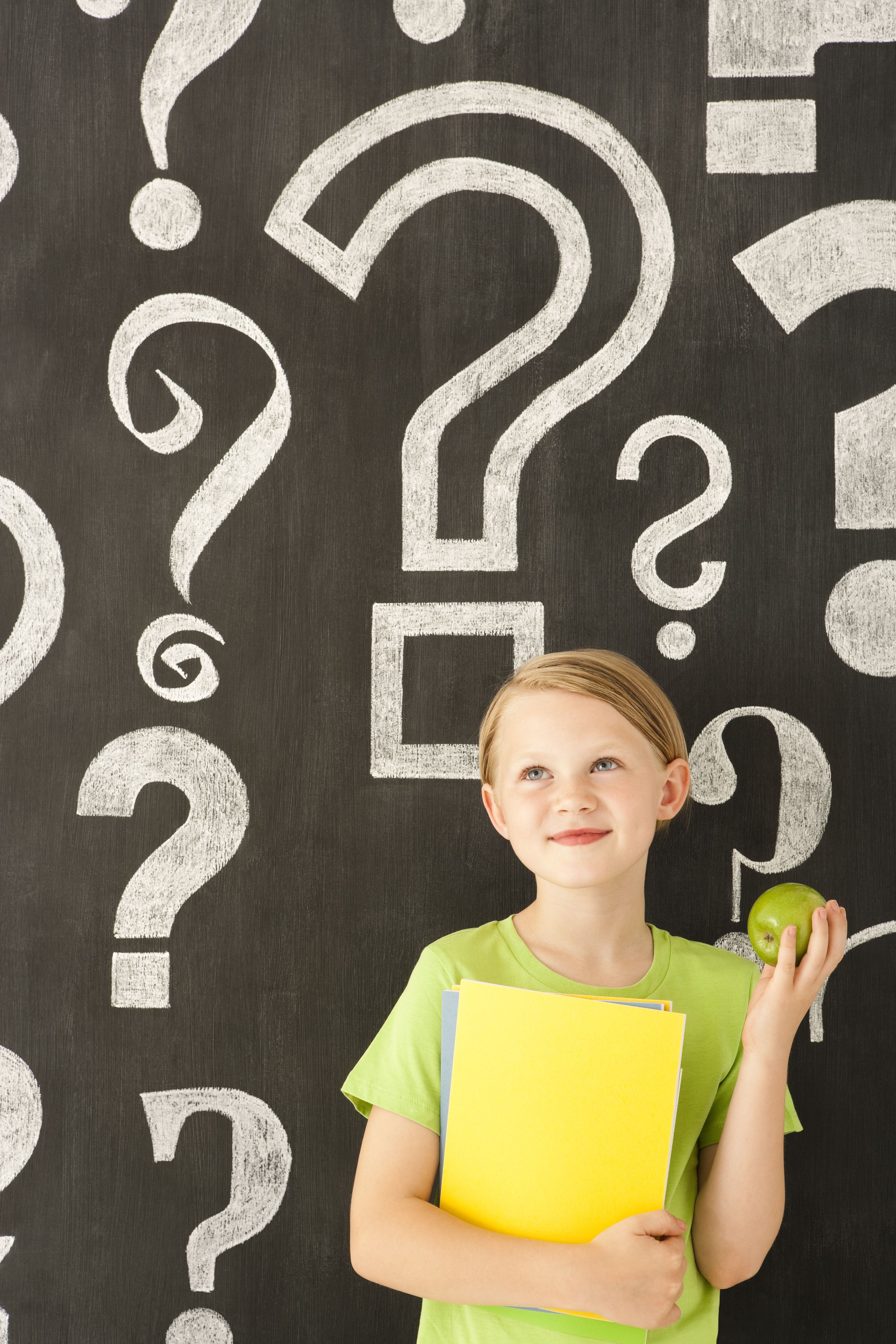 Child carrying books and an apple in front of blackboard with chalk drawn question marks